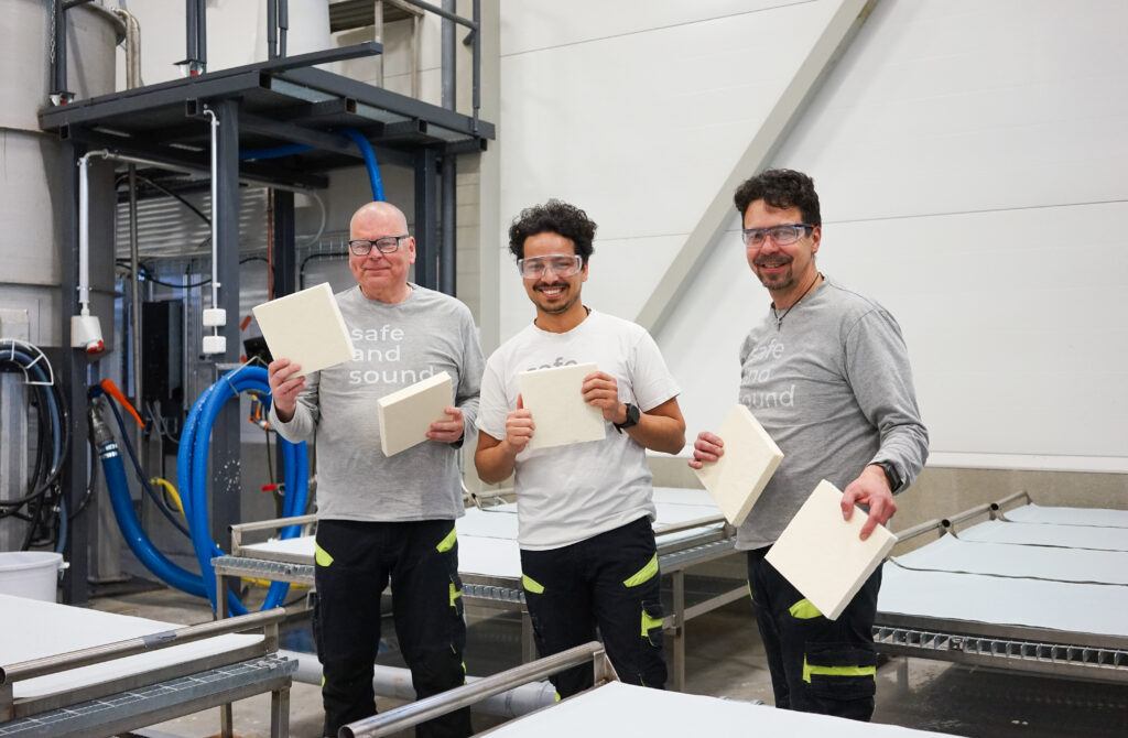 Smiling summer trainee with mentors confidently holds an acoustic tile, showcasing dedication and enthusiasm in their role.